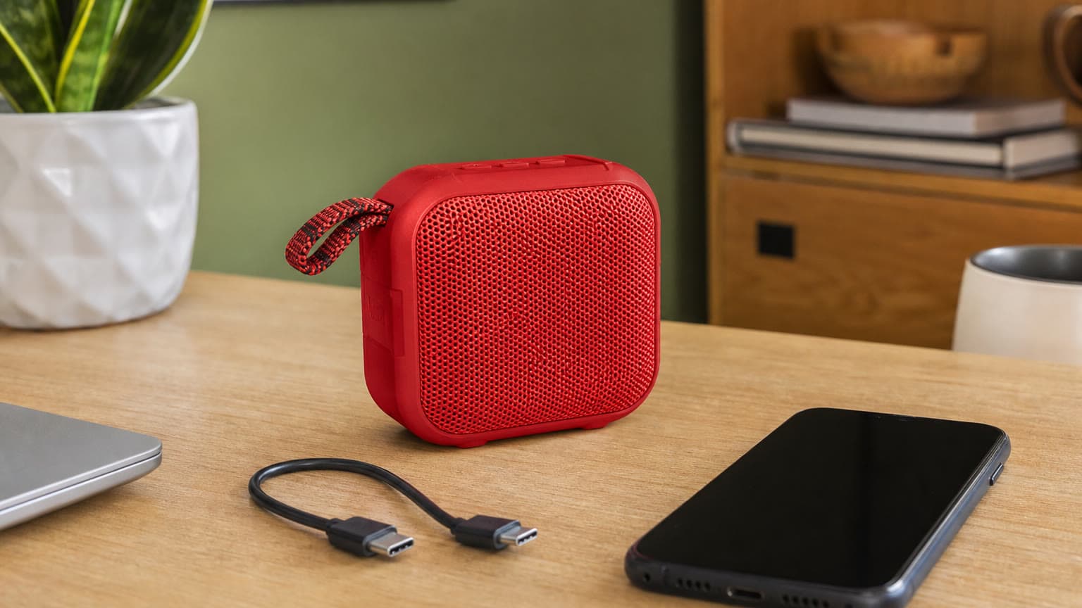 Compact red portable Bluetooth speaker on a desk with a phone and USB-C cable