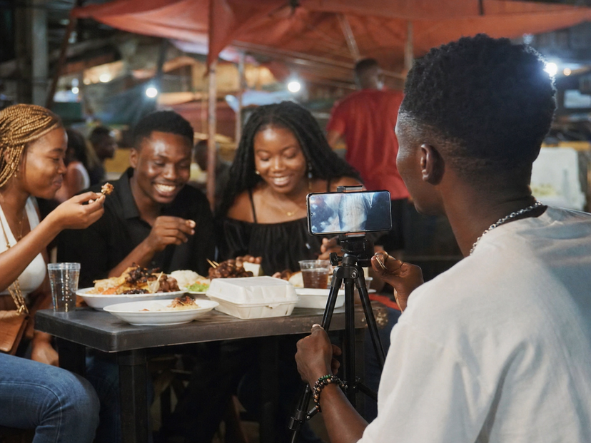 A Nigerian creator filming friends at an evening food table with a smartphone on a tripod